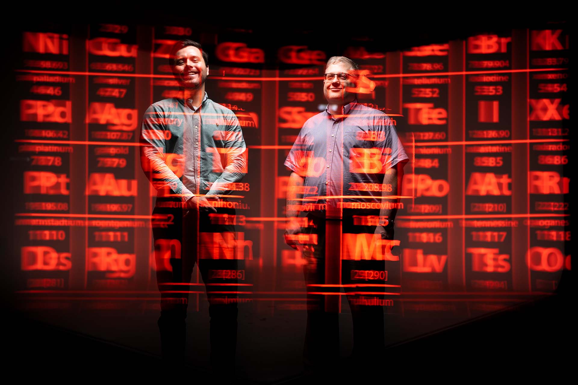 Sandia engineers stand in front of a red digital periodic table of elements.