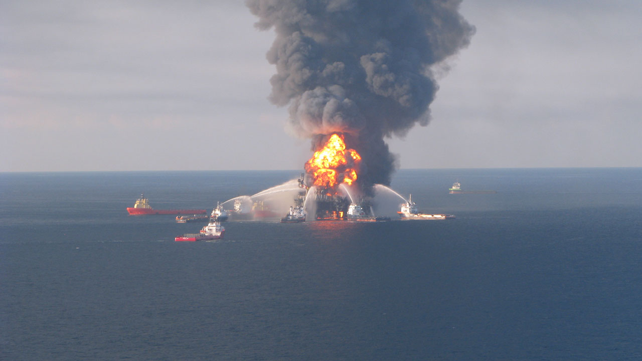 Deepwater accident. A drilling rig in the ocean is on fire. Boats are spraying water on the flames.