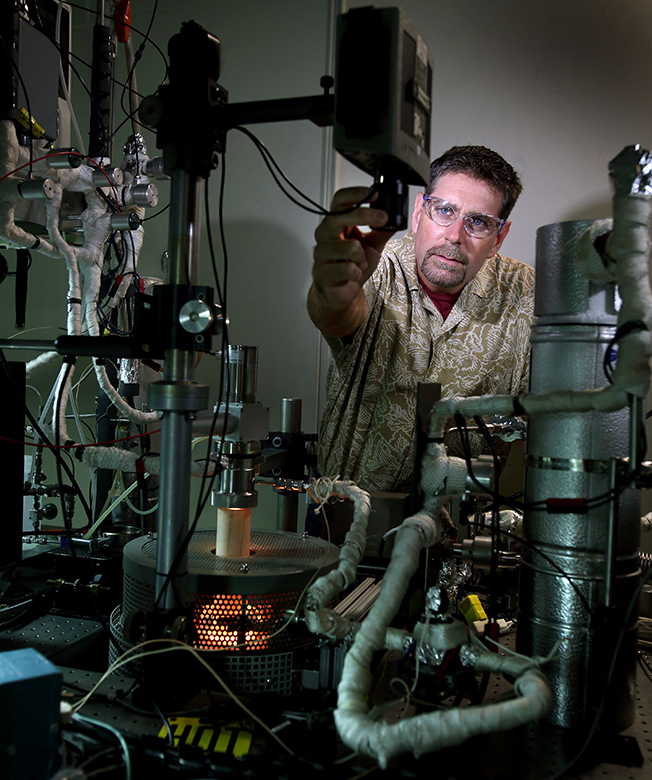 Tony McDaniel carefully adjusts the setting on an optical pyrometer used to measure the temperature of the metal-oxide particles undergoing reduction. Incandescence from the experimental reactor can be seen in the foreground and attests to the extreme temperature used to extract oxygen from the metal-oxide particles. (Photo by Dino Vournas)
