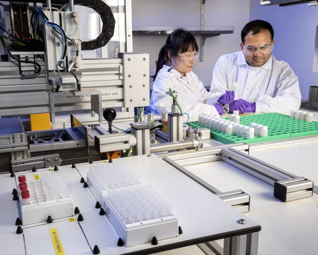 Two researchers stand behind a large, flat piece of equipment with an array of upright test tubes organized across the top