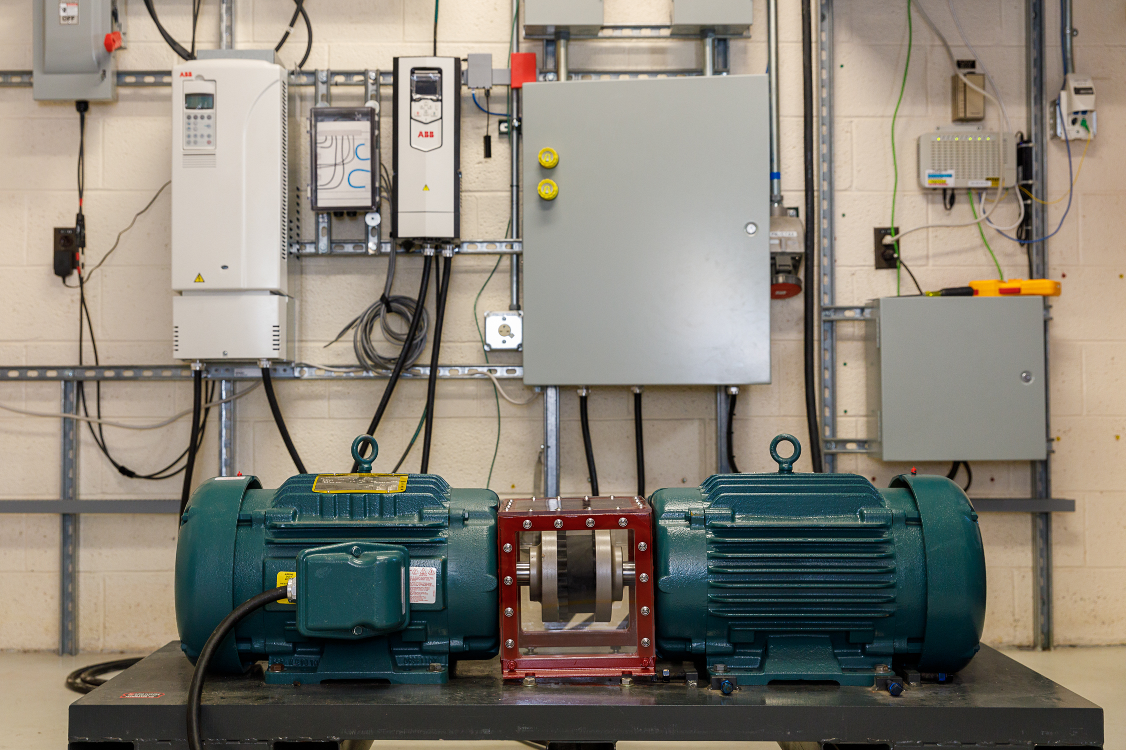 Two custom motors, cyan, are connected by a red box on a black platoform that is 4 feet wide, 2 feet deep, and 2 feet tall. Electical cables and boxes on the wall behind.