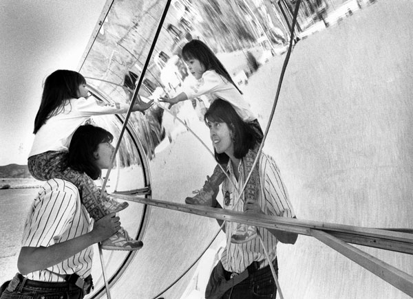 child sits on mom's shoulders as they look at their reflection in a parabolic trough