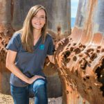 Sandia intern Rebecca Nylen kneels next to blasted steel cylinders