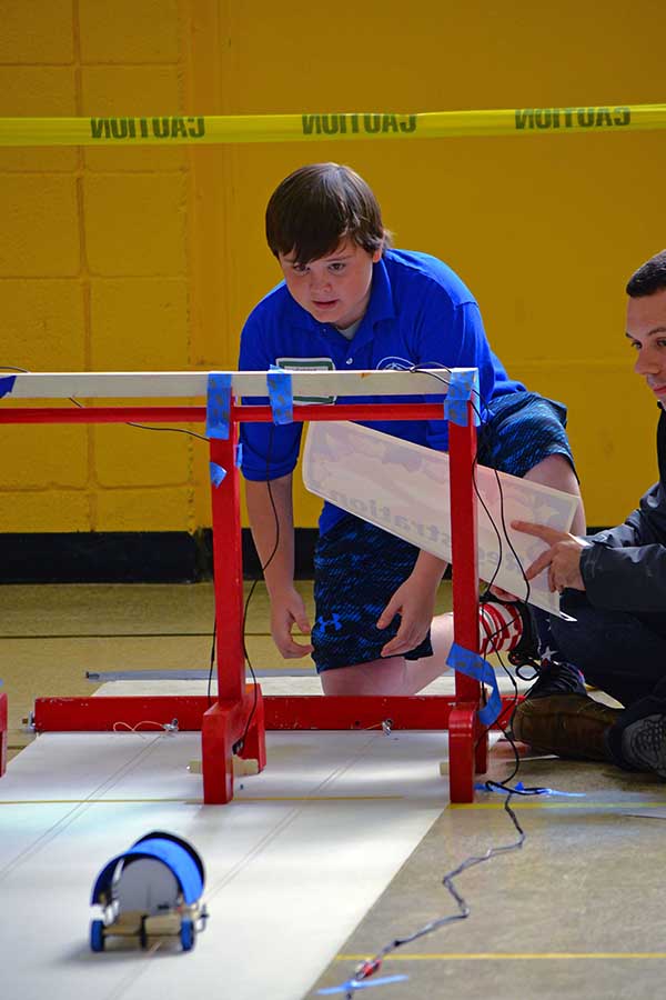 A student waits to grab his car at the end of the track while a volunteer from Sandia National Laboratories watches. More than 150 middle school students from across New Mexico participated in the challenge, presented by Sandia National Laboratories, Los Alamos National Laboratory, PNM and Intel.   The challenge, held Nov. 19 at Albuquerque’s Van Buren Middle School, is an annual STEM event in its 10th year. Students formed 5-person teams at the beginning of the school year and were provided basic materials—a lithium-ion battery, a direct-current motor, and other materials such as a chassis and wheels—needed to build their cars.  “Our goal is to expose students to basic engineering. This allows them to see how math and science intersect,” said Amy Tapia, Sandia’s community relations manager.  The teams were also required to give oral presentations on the designs of their cars, as well as the challenges they faced while constructing and running them.