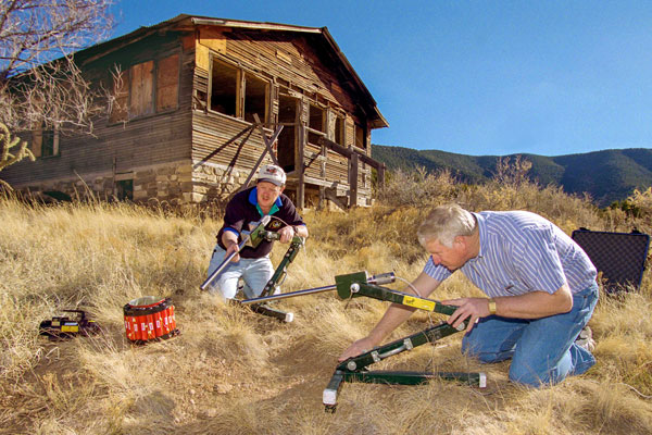 Rod Owenby and Chris Cherry use equipment on ground outside abandoned cabin