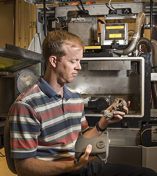 MATERIALS ASSURANCE — Bradley Jared (1832) sits in front of a new selective laser melting machine at Sandia for metal additive manufacturing (AM) as he holds two prototype housings designed through a technology called topology optimization. Sandia researchers who are exploring additive manufacturing for nuclear weapons and other national security needs say they need to understand how AM processes affect the properties of materials that are generated.	(Photo by Randy Montoya)