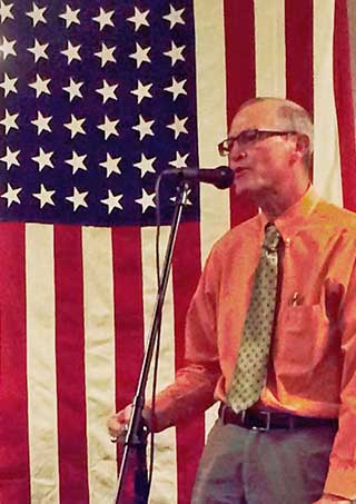 Beneath his family’s heritage US flag, Dante Berry (R) addresses the audience at a tribute to veterans in poetry, music, and art that he hosted. (Photo by Neal Singer)