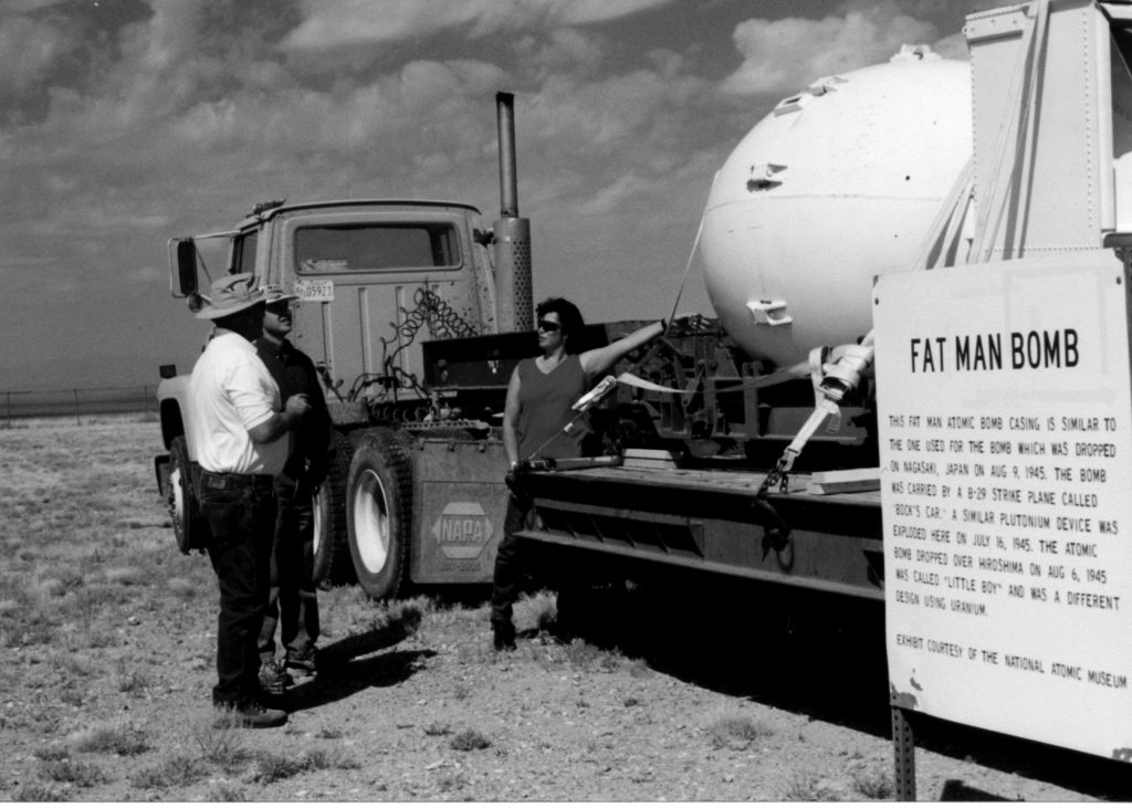 3 people standing next to a semi truck that is carrying a replica bomb