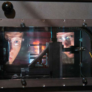 NOT SO BIG BANG — Alex Tappan (left) and Rob Knepper (both 2554) watch the detonation of a Sandia critical thickness experiment. The experiment typically uses less explosive material than the size of one-tenth of an aspirin tablet to determine small-scale detonation properties. The bench-top experiment is so small, researchers can stand next to the firing chamber with eye and ear protection. (Photo by Randy Montoya)