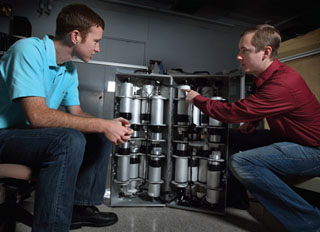 Image of <p>NNSC intern Zach Strater and his mentor Mark Gerling discuss Mobile Imager of Neutrons for Emergency Responders (MINER). While at Sandia last summer, Zach evaluated how the performance of MINER compares with conventional He-3-based detectors. (Photo by Dino Vournas)</p>