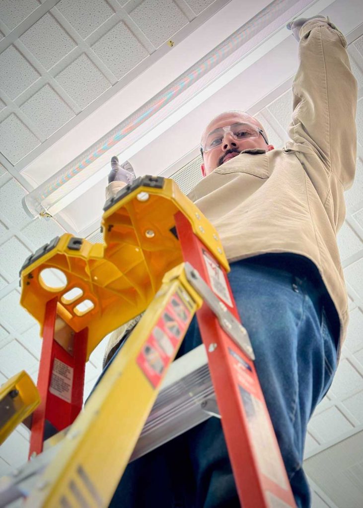 LIGHT IT UP — Lamping Crew team member Omar Rivera replaces a fluorescent bulb. This work enables staff to continue advancing Sandia’s national security mission. (Photo by James Stewart)